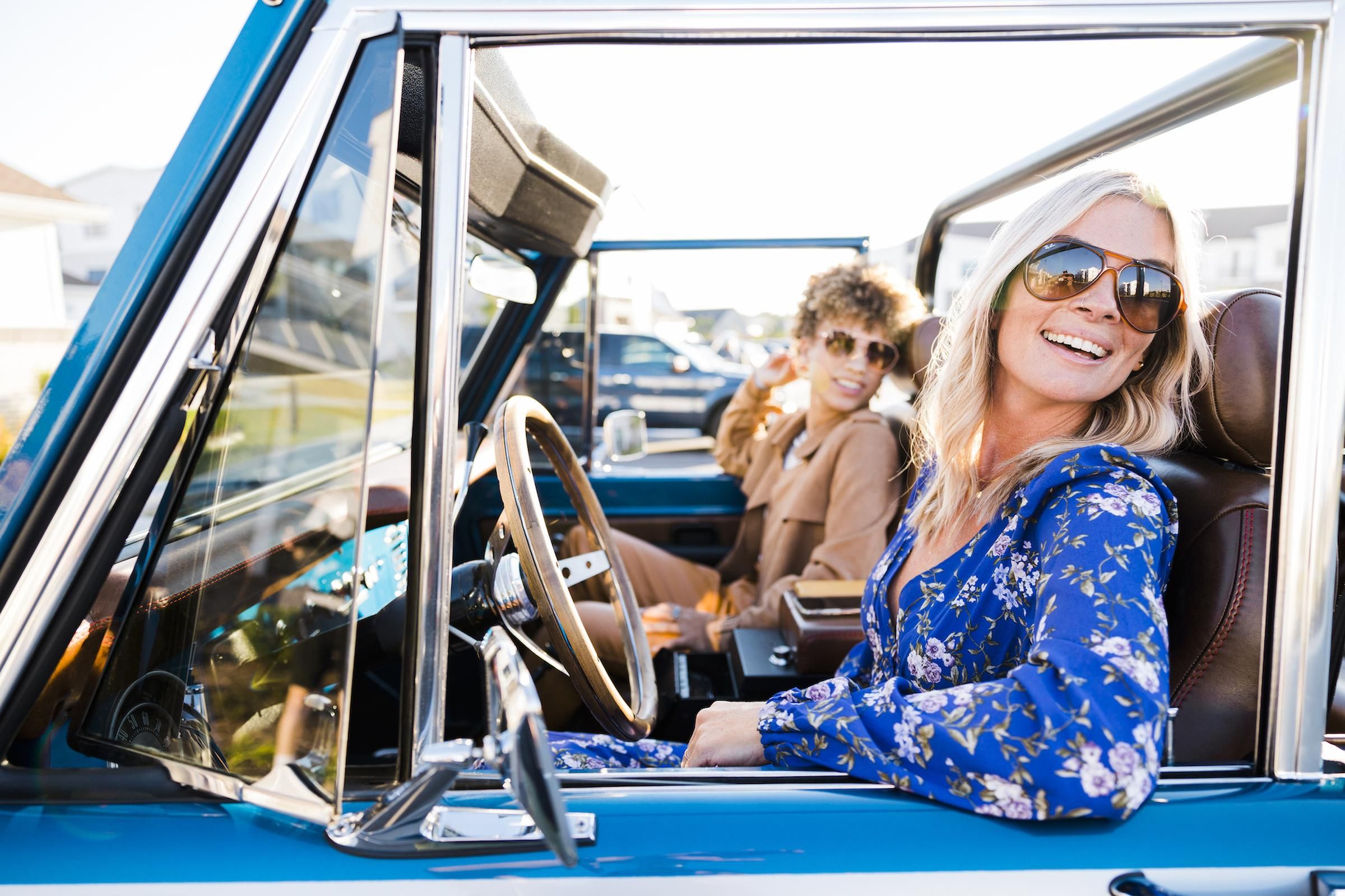 Evolve Grove Park Two women wearing sunglasses are seated in the driver's seat of a blue car, enjoying a sunny day together.
