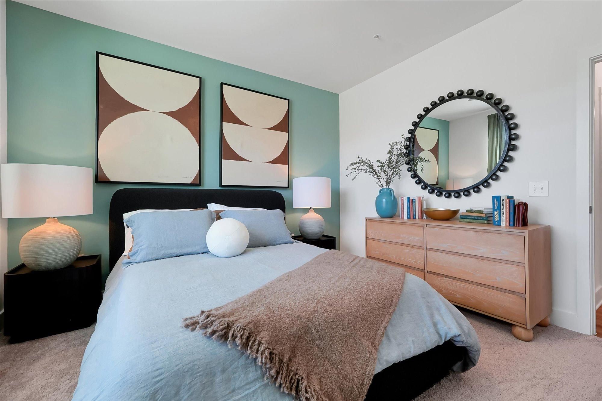 Modern bedroom with a blue accent wall, abstract art, a round mirror, and a neatly made bed with throw blanket.