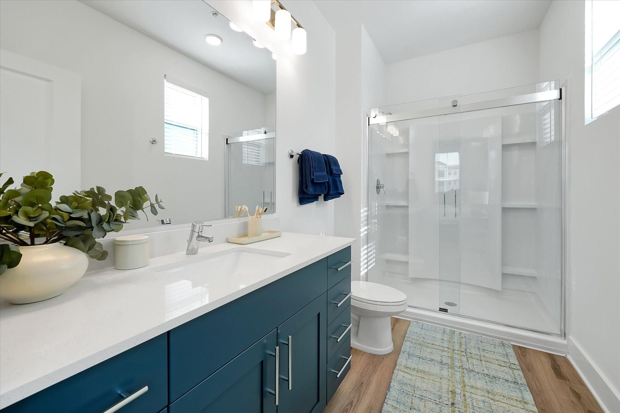 Modern bathroom with a blue vanity, white countertop, glass shower, and natural light from a window.