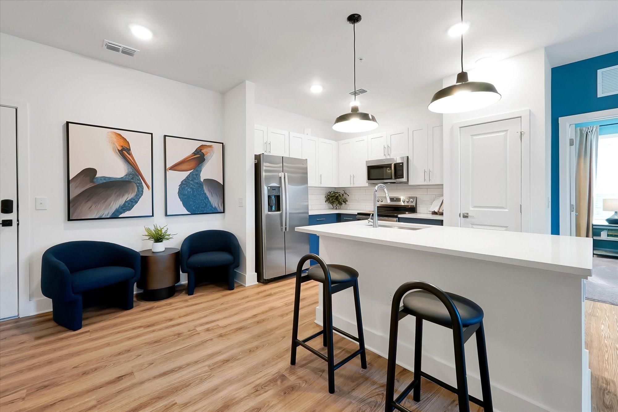 Modern kitchen with white cabinets, stainless steel appliances, bar stools, and two blue chairs by wall art.